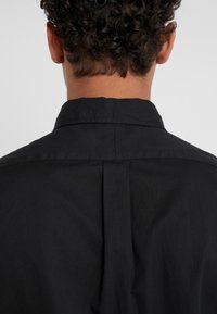 Czarna koszula z klasycznym kołnierzykiem, gładką fakturą i plisą na środku pleców, zapewniająca dopasowany krój i swobodę ruchów.