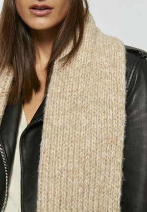 Écharpe en tricot beige faite d'un fil doux et texturé, drapée sur une veste en cuir noire, mettant en valeur son motif épais et côtelé.