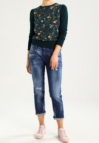 Zelená květinová mikina s dlouhými rukávy, v kombinaci s roztrhanými modrými džíny s ohrnutými manžetami a světle růžovými teniskami.