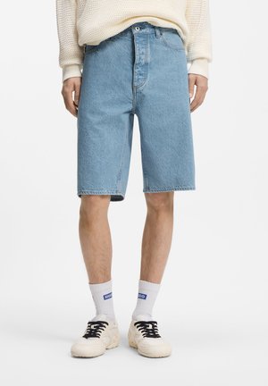 Person, die hellblaue Jeansshorts, weiße Socken mit blauem Text, weiße Turnschuhe und einen cremefarbenen Strickpullover trägt.