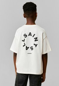 Bela bomba iz bombaža s sproščenim krojem, na hrbtni strani z okrožnim črnim logotipom z napisom "SAINT LONDON." Kratke rokave, okrogla izrezana ovratnika.