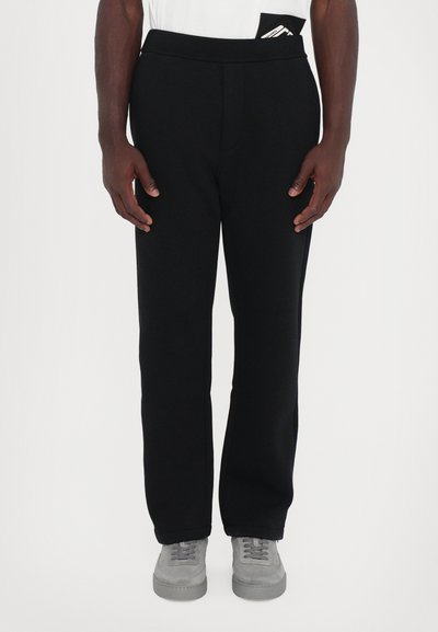 Pantalon de sport noir avec une taille élastique, jambes droites et texture lisse. Détails notables : une poche cachée dans la taille.