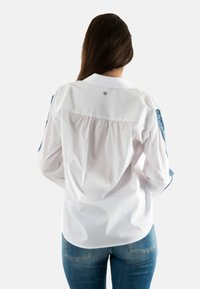 Camisa blanca de botones con la parte posterior fruncida y detalles en azul en las mangas. Hecha de una tela suave y ligera.