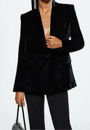 Vrouw draagt een zwarte fluwelen blazer en zwarte broek, en houdt een kleine zwarte handtas met strassversiering vast.