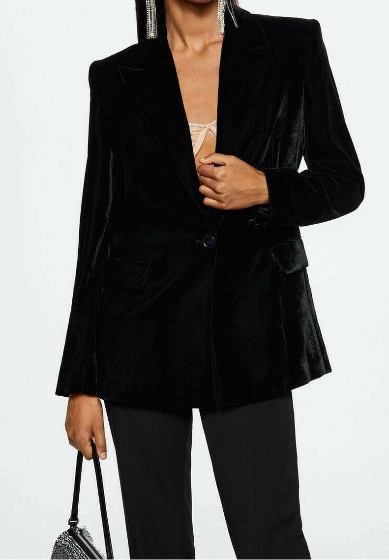 Femme portant un blazer en velours noir et un pantalon noir, tenant un petit sac à main noir orné de strass.