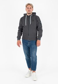 Felpa grigia con zip e cappuccio, con lacci grigi, tasche laterali e una toppa sulla manica sinistra. Abbinata a jeans blu e sneaker bianche.