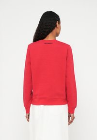 Személy göndör, sötét hajú, piros hosszú ujjú pulóvert visel, melynek a nyakvonala közelében "KARL LAGERFELD" felirat olvasható, valamint fehér szoknyát.