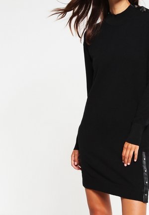 Femme portant une robe longue noire en tricot avec des détails de boutons sur l'épaule et le long d'un côté près de l'ourlet.