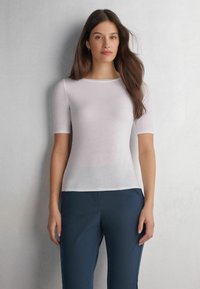 Hvid kortærmet bluse lavet af gennemsigtigt stof med rund halsudskæring, parret med mørkeblå bukser. Glat tekstur, minimal design.