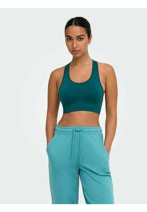Femme aux cheveux foncés portant un débardeur court bleu sarcelle et un pantalon de jogging turquoise, debout les mains dans les poches sur un fond uni.
