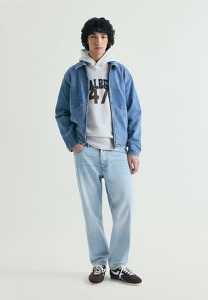 Joven de pie con las manos en los bolsillos, vestido con jeans azul claro, sudadera gris con "Calpe 47", chaqueta vaquera azul y zapatillas negras.