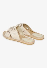 Sandalias planas beige con tiras cruzadas, elaboradas con tela suave. Suela de yute natural, plantilla lisa con detalle de logo y diseño plano.