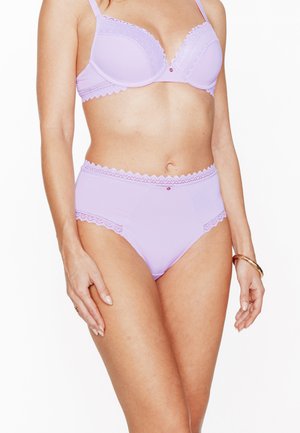 Femme portant un ensemble soutien-gorge avec bordure en dentelle lavande et culotte taille haute, debout devant un fond blanc uni.