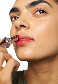 Red lipstick in a silver and pink tube being applied to a model’s lips. Smooth, glossy texture; model has clear skin and dark brows.