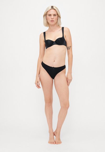 Mulher com cabelo loiro curto, vestindo um soutien e uma calcinha pretos, de pé descalça diante de um fundo branco liso, com uma tatuagem de borboleta no braço esquerdo.