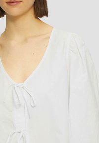 Weiße Bluse mit V-Ausschnitt, Schleifenverschluss vorne und puffärmel. Hergestellt aus einem strukturierten Stoff und zeigt ein sauberes, minimalistisches Design.