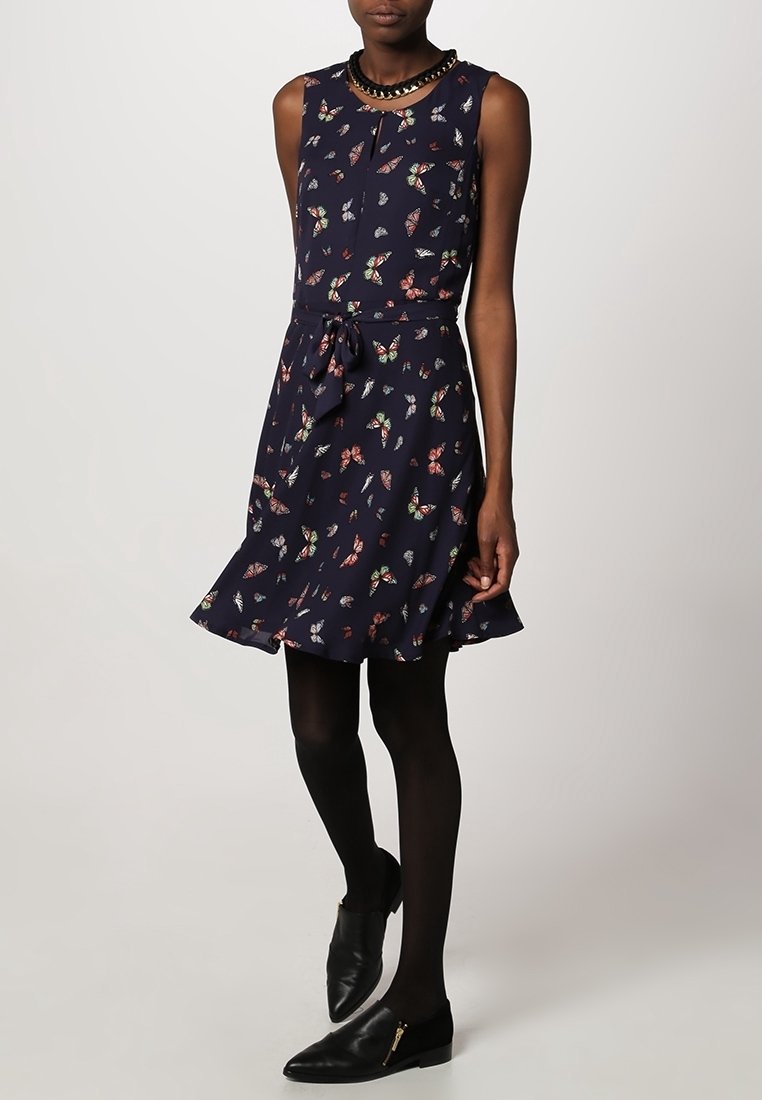 Femme portant une robe sans manches bleu marine ornée d'un imprimé papillons colorés, des collants noirs, des mocassins en cuir noir et un collier noir en perles.