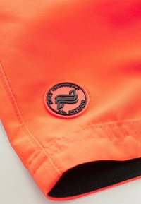 Kirkkaan oranssit uima-asu-shortsit, joissa on musta kuminen logo-patch, jossa lukee "NEXT SWIMWEAR" ja "No. 987248" helmassa. Sileä rakenne.