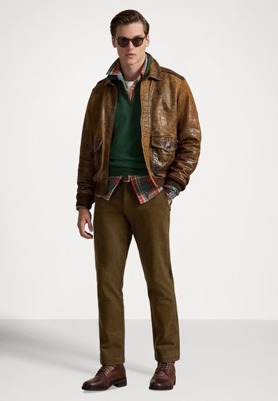 Καφέ δερμάτινο μπουφάν bomber, πράσινο πουλόβερ, πουκάμισο καρό, παντελόνι χακί, σκούρα καφέ μπότες. Τα γυαλιά ηλίου ολοκληρώνουν το σύνολο.