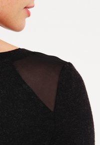 Svart tröja med en texturerad stickad kropp och en genomskinlig svart panel på axeln. N ärbild på halsringningen och axeldetaljen.