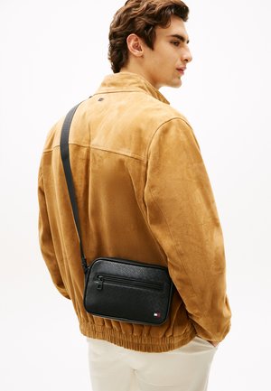 Jonge man die een lichtbruine suède jas en witte broek draagt, met een zwarte crossbodytas met een textuurpatroon en een voorvak met ritssluiting.