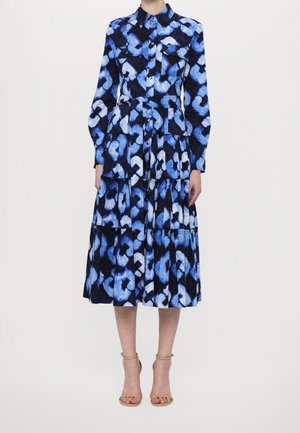Femme portant une robe midi à manches longues, boutonnée, bleu et noir à motifs, avec col et jupe étagée, associée à des sandales beiges à talons hauts.