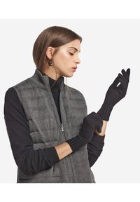 Svarta stickade handskar med ribbade manschetter, bärs av en modell i en grå väst över en svart topp, vilket visar upp värme och mångsidighet i designen.