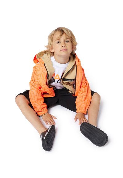 Heldere oranje hooded jas met beige accenten, voorzien van een logo-opdruk en ritsluiting. Zwarte shorts en instap schoenen maken de outfit compleet.