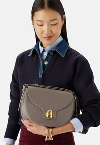 Femme aux longs cheveux foncés portant une chemise à couches et un pull, tenant un sac à main en cuir gris avec un fermoir doré et portant des bracelets dorés.