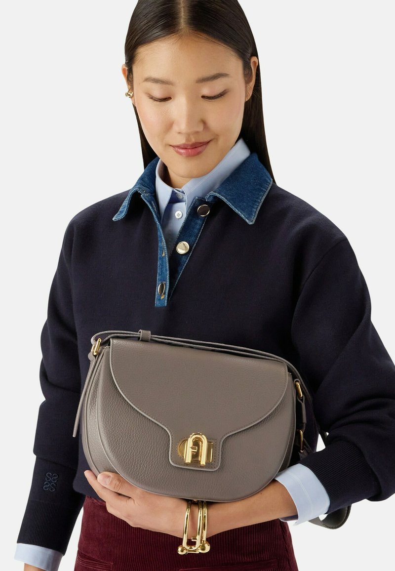 Femme aux longs cheveux foncés portant une chemise à couches et un pull, tenant un sac à main en cuir gris avec un fermoir doré et portant des bracelets dorés.