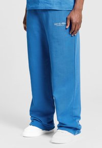 Blå bomulds-sweatpants med en afslappet pasform, med sidelommer og broderet logo på venstre lår, parret med hvide sneakers.