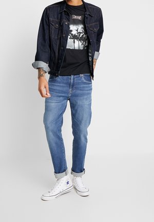 Man draagt een donker denim jack over een zwart T-shirt met palmboomprint, blauwe opgerolde spijkerbroek en witte hoge sneakers tegen een witte achtergrond.