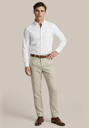 Man draagt een wit overhemd met knopen, beige geplooide broek met een bruine riem en bruine leren nette schoenen, staand met hand op de heup.