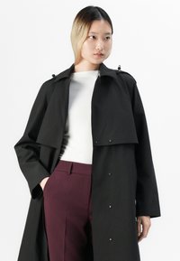 Giovane vestito con un trench nero, una maglietta bianca e pantaloni bordeaux, con una mano in tasca, che guarda leggermente di lato.