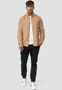 Camisa de botones color marrón claro sobre una camiseta blanca, combinada con pantalones negros ajustados y zapatillas blancas con detalles en negro.