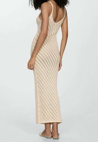 Donna con capelli ricci che indossa un vestito maxi senza maniche di maglia beige e scarpe slip-on piatte beige, in piedi con la schiena rivolta in avanti.