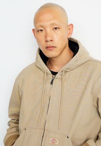Tan hooded jacket with a zipper, featuring a front pocket and drawstrings. Worn over a black shirt, with visible stitching and logo detail.