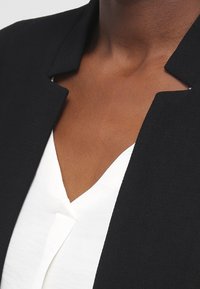 Blazer noir avec un col structuré, associé à un chemisier blanc à col en V. Texture de tissu lisse et lignes épurées sans ornements.