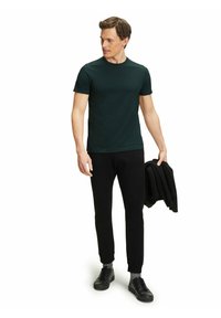 T-shirt verde scuro a maniche corte abbinato a pantaloni neri a sigaretta e sneaker nere. Il modello tiene in mano una giacca nera piegata. Texture liscia.