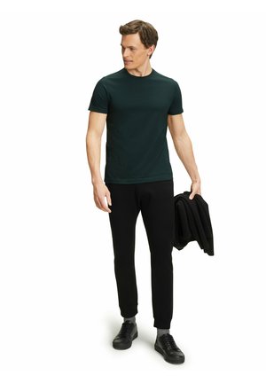 Dunkelgrünes, kurzärmliges T-Shirt, kombiniert mit schwarzen, enganliegenden Hosen und schwarzen Sneakern. Das Model hält eine gefaltete schwarze Jacke. Glatte Textur.