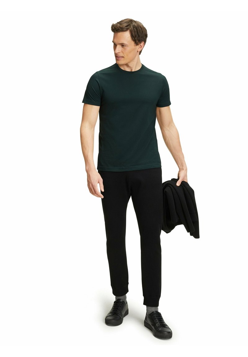 T-shirt verde scuro a maniche corte abbinato a pantaloni neri a sigaretta e sneaker nere. Il modello tiene in mano una giacca nera piegata. Texture liscia.