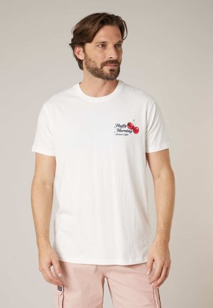 Homme avec barbe portant un t-shirt blanc avec le texte "Fluffy Morning" et un graphisme de cerise sur la poitrine, associé à un short cargo rose clair.