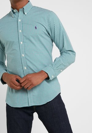 Homme portant une chemise à petits carreaux verts et blancs boutonnée avec un logo brodé violet, et un jean bleu foncé, les mains jointes à la taille.