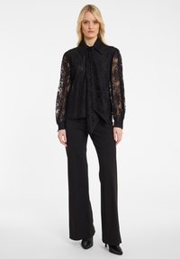 Schwarze Spitzenbluse mit hohem Kragen und Schleife, über einem einfarbigen schwarzen Shirt getragen. Kombiniert mit schwarzen, ausgestellten Hosen mit subtilen Verzierungen.