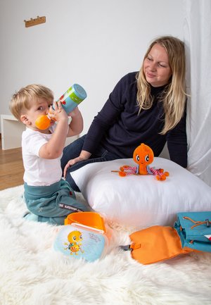Colorful plastic sippy cup, plush orange octopus toy, blue and orange bags, and a white pillow on a soft white rug. Wooden wall hook visible.