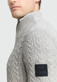 Pull en laine gris clair avec texture en maille torsadée, col montant et étiquette de marque noire sur la manche. Présente un design ajusté et des accents côtelés.