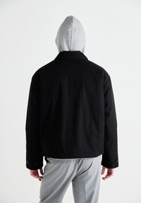 Black jacket with a collar, featuring a grey hood. Composed of a smooth fabric, displaying button accents on the sleeves, worn over grey trousers.