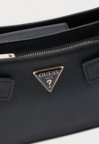 Μαύρη υφασμάτινη τσάντα με χρυσές λεπτομέρειες, που διαθέτει τριγωνική πλακέτα με λογότυπο. Άνοιγμα με φερμουάρ, δομημένο σχήμα και διπλές λαβές.