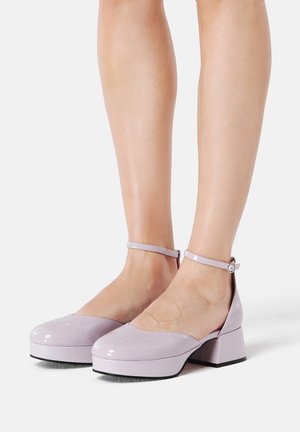 Chaussures à talons en cuir verni lilas avec un bout rond, une bride à la cheville et un talon bloc. Texture lisse et design minimaliste.