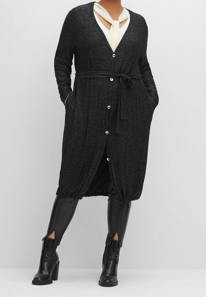 Femme portant un long cardigan gris foncé boutonné avec ceinture nouée, blouse blanche, leggings en cuir noir et bottes à talons hauts noires.